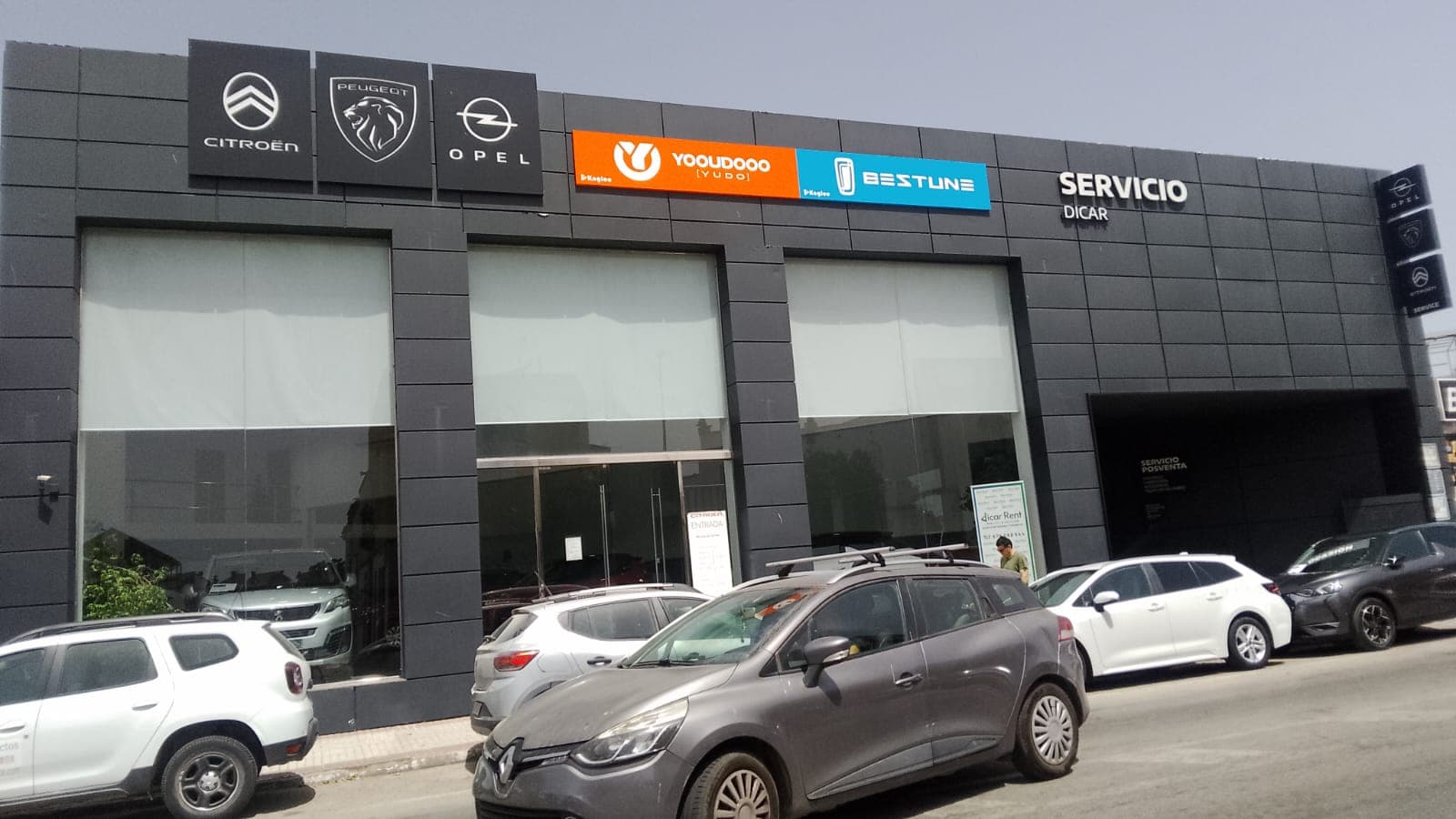 Fachada de concesionario con logotipos de Citroën, Peugeot y Opel y varios coches aparcados delante.